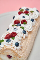 Mixed berry pavlova roll with cream and mint on white plate with pink background