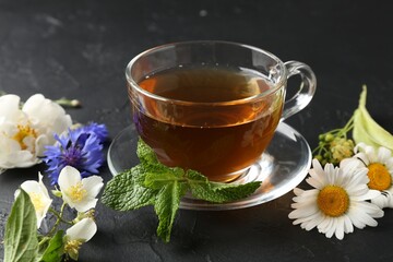 Aromatic herbal tea in glass cup, different flowers and mint on black table, closeup