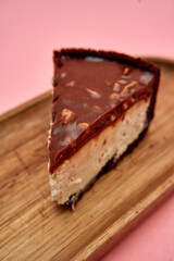 Chocolate-covered cheesecake slice on wooden plate with nuts highlighted against pink background