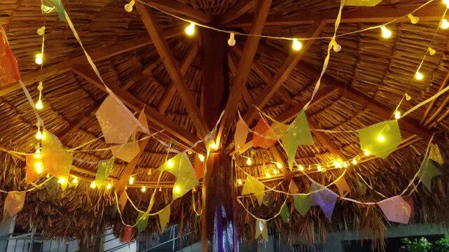Looking up at a rustic thatched roof decorated with colorful festive pennant flags and warm string lights. Cozy nighttime atmosphere of a traditional party, June celebration, fair or gathering.