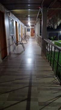 Peaceful night view of an illuminated outdoor corridor at a rustic tropical resort. Empty chairs line the long hallway with tiled floors, leading to guest rooms under a thatched roof.