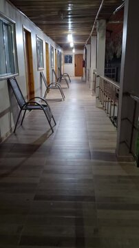  Walking view of peaceful hotel veranda at night with empty chairs along a rustic wall, warm lights illuminating the corridor, and a view of the garden. Cozy and tranquil vacation resort architecture.
