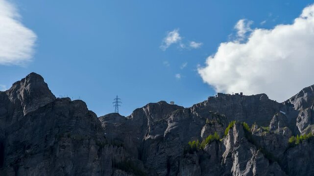 Mountain peak in Leukerbad named Gemmi. The cable car ascends the mountain. Power poles are visible. Time-lapse of clouds in the sky. The natural landscape is in bloom. Tourist area.