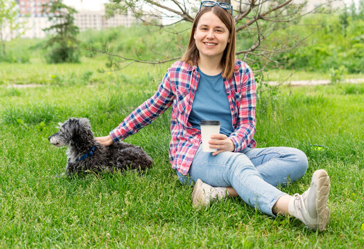 Happy smiling young woman sitting on grass with mixed breed gray fluffy senior dog bedlington terrier whippet, enjoying coffee in a park, relaxed atmosphere and joyful moment captured in nature