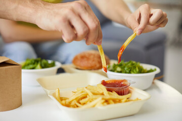 Man dipping French fries into ketchup to eat potato sticks with spicy red sauce at home. Fingers of hungry guy take chips out of open package to dip into catsup. Fast food restaurant delivery concept