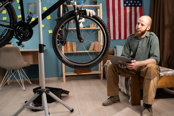 Bearded mechanic doing bike servicing using laptop at home searching replacement components online....