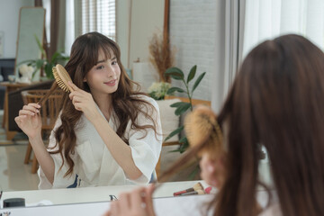 Teenage multiracial woman combing hair gently in front of mirror during morning grooming routine natural soft light scene bathrobe makeup desk beauty and skincare self care concept