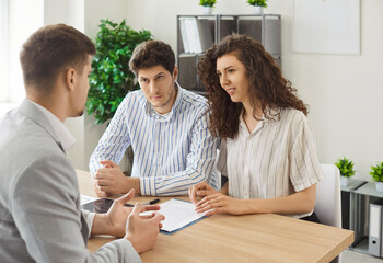 Young family couple sitting at desk and listening to male real estate agent in office. Focused...