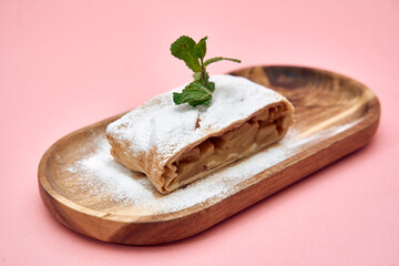 Apple strudel slice on wooden plate with mint leaf and powdered sugar on pink background