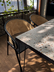 An indoor-outdoor cafe setting featuring a terrazzo table, rattan chairs, and natural light with climbing plants in the background.