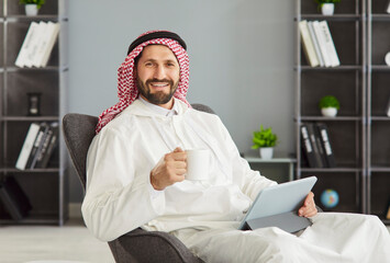 Happy Arabian man having coffee break during doing remote work, posing with cup of coffee and...