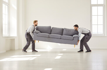 Two movers in uniform lifting large sofa and bringing it into bright empty room. Professional...