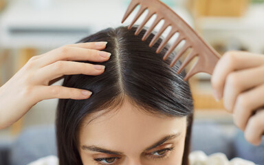 Fototapeta premium Closeup of brunette young woman brushing her graying head with a comb indoors. Female person examining her scalp and black hair looking at gray hairs at the roots standing at home.