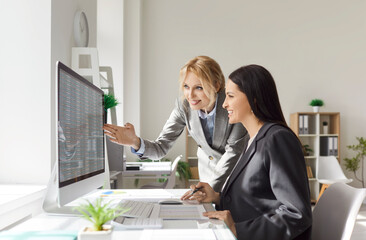 Two successful women analyzing financial details and looking through digital file on PC, posing at...