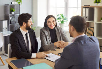Happy smiling couple shaking hands reaching agreement with a man realtor or financial advisor signing successful contract sitting at desk on workplace. Insurance agent consulting clients in office.