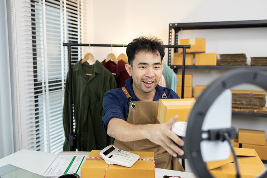 A man is live streaming his shirt on his mobile phone. On the table are boxes and shipping supplies. He is wearing a blue shirt and a brown apron.

