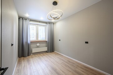 Bright, empty room with wood floors, grey walls, and a window with grey curtains. A modern, circular chandelier hangs from the ceiling