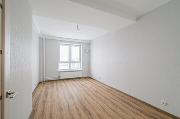 empty, bright room with wood-like flooring. Walls are textured white/gray. A large window faces a cityscape. A partially-seen door is on the left