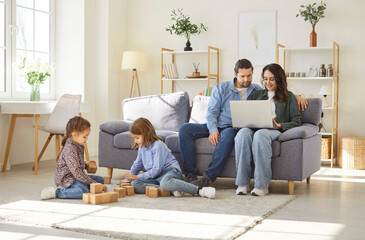 Family at home, active daughter and son playing at floor with cubes, parents sit on sofa resting...