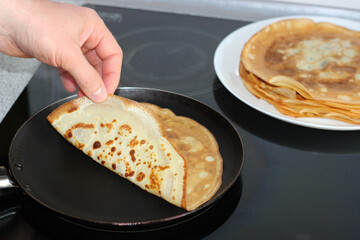 Man flipping homemade crepes on frying pan using a wooden spatula and fingers. Cooking thin pancakes in a cozy home kitchen. Rustic breakfast in progress, close-up, side view