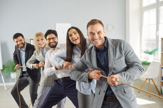 Cheerful multicultural business team playing tug of war together in office. Smiling colleagues pulling rope during teamwork activity, showing unity, energy, motivation and collaboration spirit.