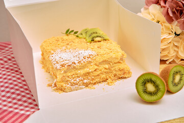 Delicious napoleon cake in box with fresh kiwi and flowers on checkered tablecloth