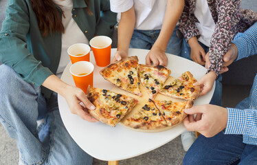 Cropped top view of family members reaching for slices of hot pizza on wooden plate over white table with orange paper cups nearby. Parents and children enjoying delicious food together during dinner.