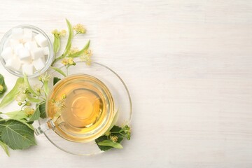 Fresh linden tea in cup, sugar cubes and flowers on white wooden table, flat lay. Space for text