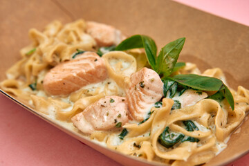 Creamy salmon tagliatelle with spinach and basil in cardboard tray on pink background