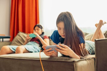 Asian half-breed girl on sofa smiling, playing on smartphone, headphones, orange charging cable connected, blurred background, sister, friend, technology education entertainment