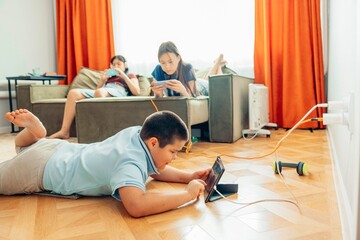 boy lying on wooden floor playing with tablet while two girls relax on sofa using smartphones with orange curtains, technology education leisure, power outlet with charging cables