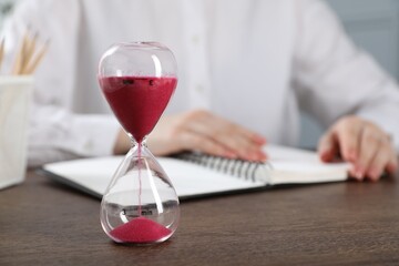 Deadline. Woman with notebook working at wooden table in office, focus on hourglass