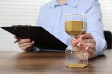 Deadline. Woman with hourglass and clipboard at wooden table in office, closeup