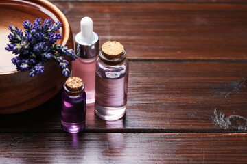 Bottles of lavender essential oil, flowers and bowl with water on wooden table, closeup. Space for text