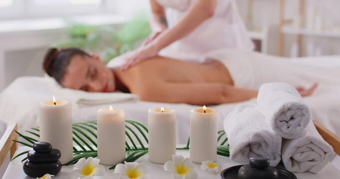 Young woman feeling happy and relax during back massage lying on couch with closed eyes in spa beauty salon with candles in foreground. Skillful masseuse massaging client. Wellness concept. 4k video. - Powered by Adobe