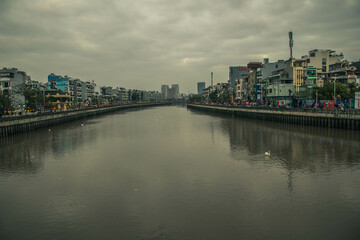 The Tam Bac riverside promenade is alive with visitors enjoying the cool evening. City buildings line the bank, reflecting off the calm water as dusk approaches in Hai Phong city, Vietnam