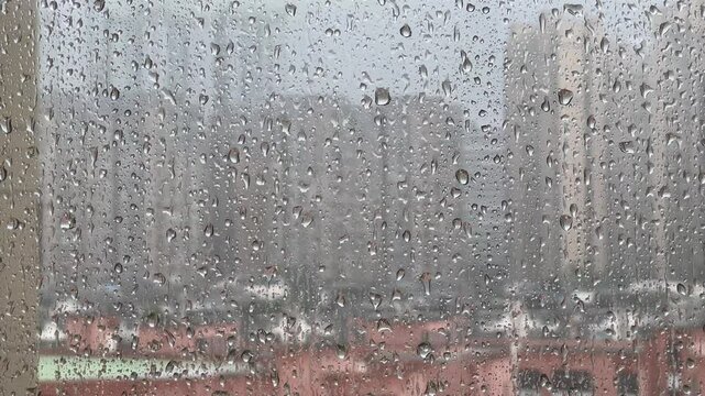 Rainy day scene: Raindrops on the window. Raindrops slide down a glass window. Outside, city buildings are blurred by the rain. The sound of rain complements the tranquil, wet atmosphere.