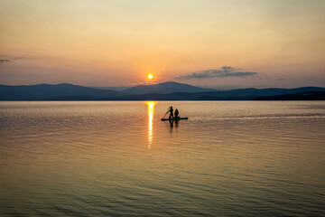 Silhouette of a girl and a child on a SUP board in the rays of the setting sun on a picturesque lake