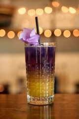 Purple and yellow layered cocktail with flower garnish in glass with straw on bar counter