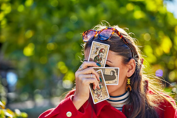 A woman in a red coat holds tarot cards in front of her face while sitting outdoors in Montoro,...