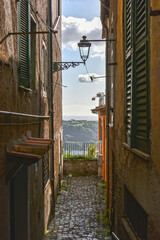 narrow cobblestone alleyway with vintage shutters and lanterns opens to a scenic view over hills and a lake. The warm afternoon light enhances the charm of this picturesque village.