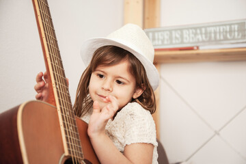 little girl wearing a white hat gently touches the strings of an acoustic guitar at home. A tender moment of childhood curiosity and musical discovery. Generation alpha concept.