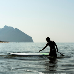 Man walking with a paddle next to his board in shallow water with mountain view in the background. Silhouette scene on a calm sea during a sunny day. Outdoor sport and lifestyle concept.