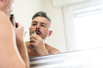 Man trimming his beard and mustache with an electric razor while looking at himself in the bathroom mirror. A moment of male personal care and grooming routine at home. Daily hygiene.