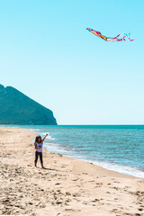 young girl flying a colorful kite on a deserted beach with clear blue sky and mountain in the background. A joyful moment of freedom and childhood at the seaside. Gen alpha and outdoor play concept.