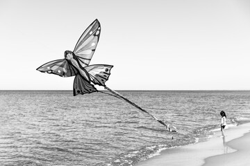 large butterfly kite flying above the sea while a young girl stands quietly at the shoreline. A poetic and nostalgic scene of childhood captured in black and white. Solitude and gen alpha concept.