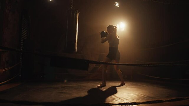 Cinematic slow motion shot with beautiful athletic lady punching bag in box ring . Physical exercise for keeping fit and stay beautiful in middle age, practicing arms punches, female boxer fighter