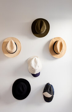 Six hats being mounted on smooth white wall in staggered rows, featuring straw fedoras and caps