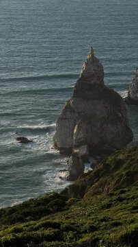 Sunlight illuminates a dramatic, pointed rock formation rising from the ocean as gentle waves break around its base, creating a mesmerizing coastal landscape. Portugal. Caba de roca. Vertical video