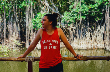 A woman leans against a wooden railing, enjoying the serene mangrove trees, calm water, and natural scenery in casual attire. The environment exudes leisure and connection with nature.